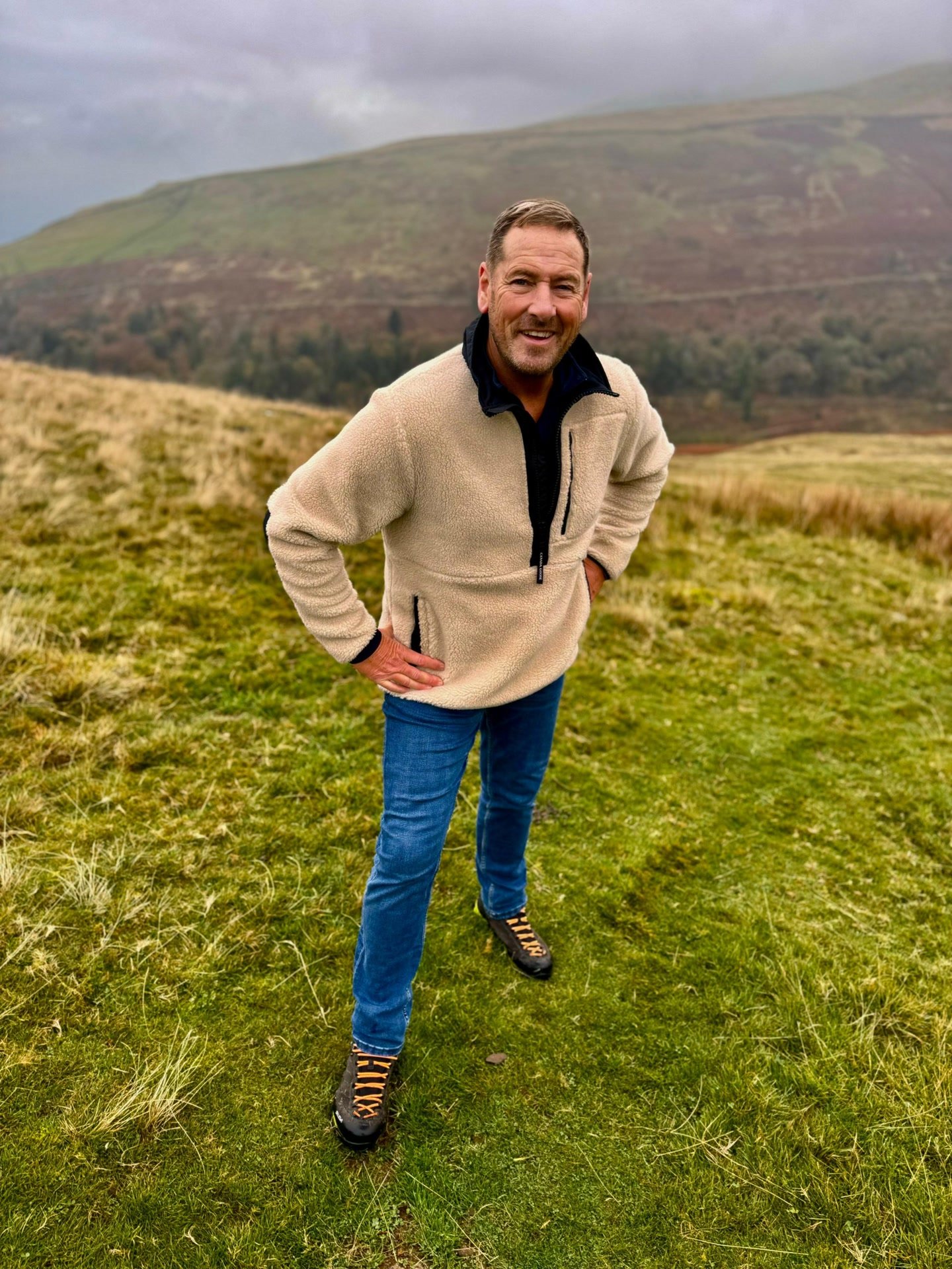 James standing on a green hillside in a cream fleece, smiling, with a moorland ridge behind him