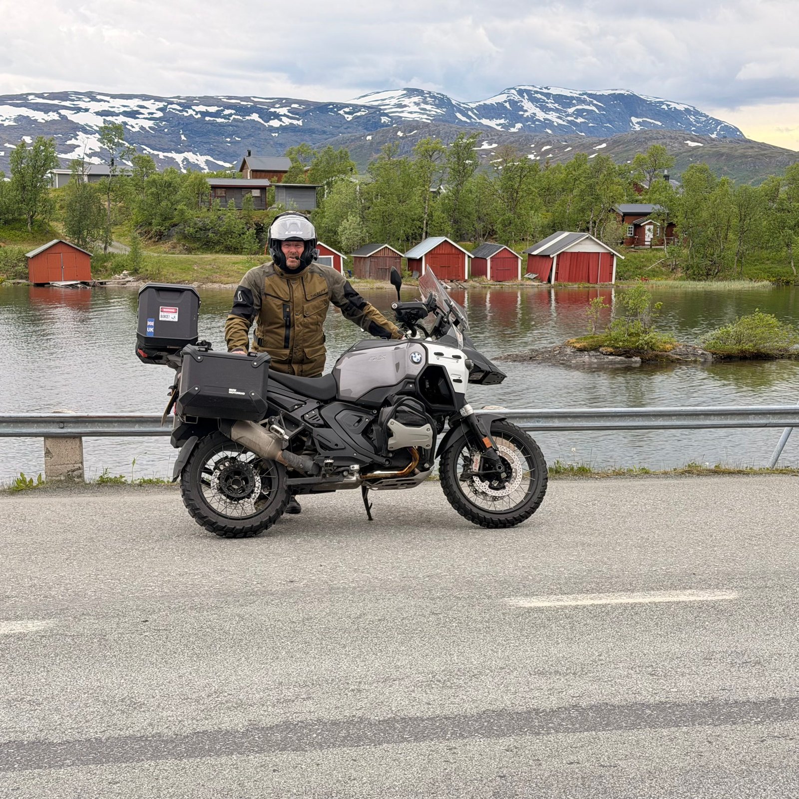 James in motorbike gear beside his BMW GS by a Norwegian lake with red boathouses and snowy mountains